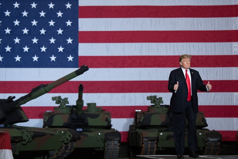 Donald Trump does two thumbs ups in front of a giant American flag and several tanks