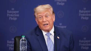 Donald Trump speaks while seated at a table with a "Ratepayer Protection Pledge" backdrop behind him.
