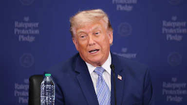 Donald Trump speaks while seated at a table with a "Ratepayer Protection Pledge" backdrop behind him.