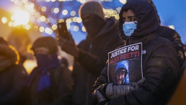 A woman wearing a face masks and a winter coat holds a piece of paper that reads "Justice for Alex Pretti" with a photo of his face. Others stand near her, also wearing face masks.