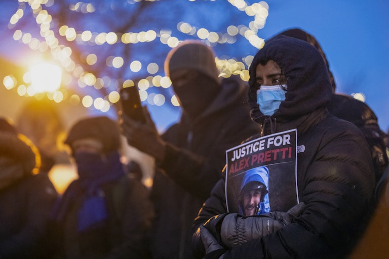 A woman wearing a face mask and a winter coat holds a piece of paper that reads "Justice for Alex Pretti" with a photo of his face. Others stand near her, also wearing face masks.