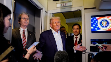 Donald Trump shrugs aboard Air Force One, as Scott Bessent and Marco Rubio stand on either side of him. Reporters surround them, holding their phones out to record.