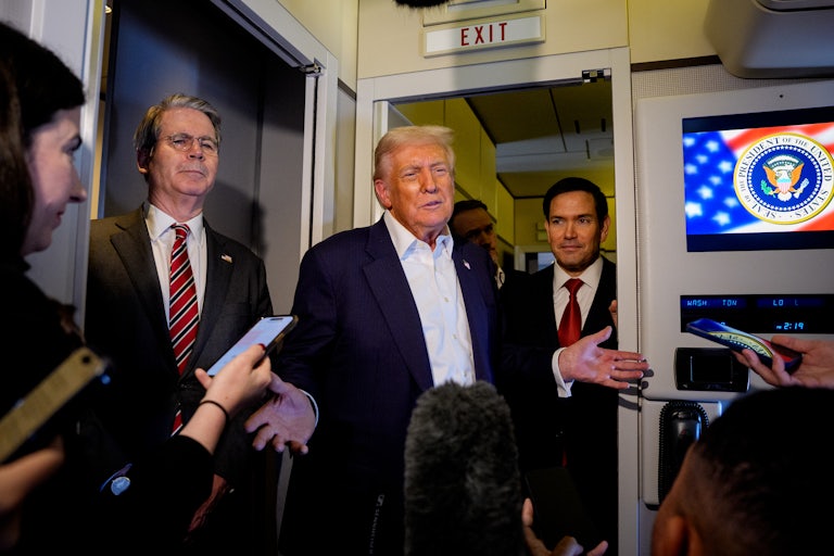 Donald Trump shrugs aboard Air Force One, as Scott Bessent and Marco Rubio stand on either side of him. Reporters surround them, holding their phones out to record.