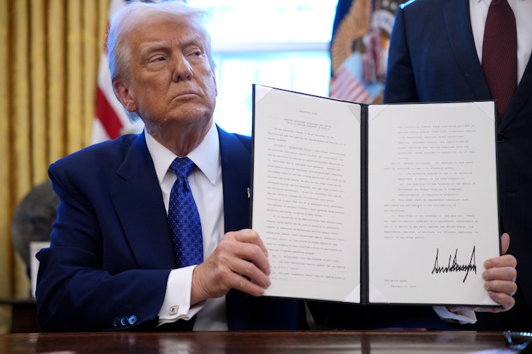 Donald Trump holds up a signed executive order while sitting at his desk in the Oval Office.