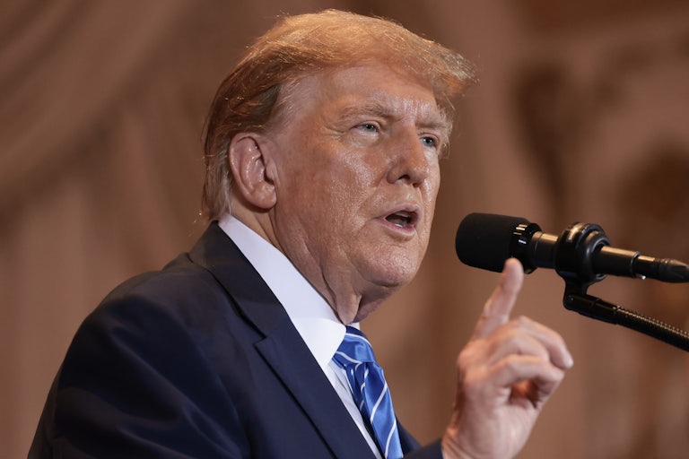 Donald Trump speaks at a mic and holds up his right index finger for emphasis. You can see the outline of his spray tan on his face (it's bad).