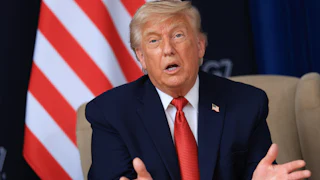 Trump speaks with his hands and mouth open on a beige chair, with the stripes of an American flag visible behind him. He's wearing a blue suit and red tie with a American flag lapel pin.