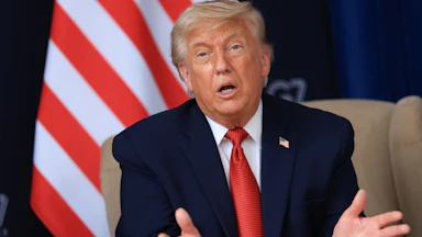 Trump speaks with his hands and mouth open on a beige chair, with the stripes of an American flag visible behind him. He's wearing a blue suit and red tie with a American flag lapel pin.