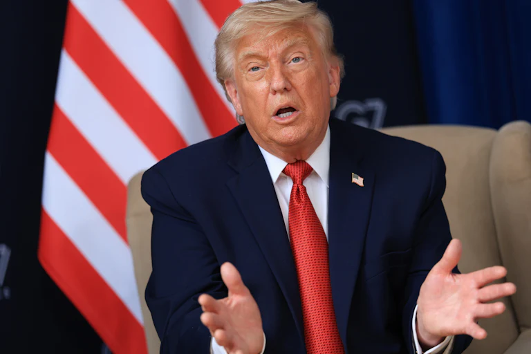 Trump speaks with his hands and mouth open on a beige chair, with the stripes of an American flag visible behind him. He's wearing a blue suit and red tie with a American flag lapel pin.