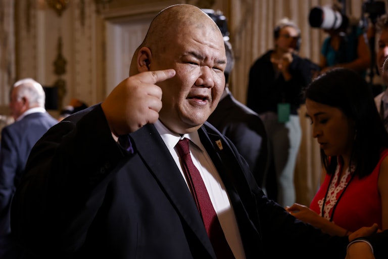 Trump spokesman Steven Cheung speaks and points a finger to his head. A press camera and other people (out of focus) are in the background.