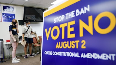 A sign in the forefront reads "Stop the Ban Vote No August 2 on the constitutional amendment." Two people stand in the background wearing masks.