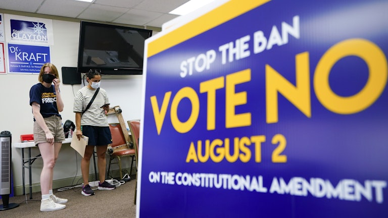 A sign in the forefront reads "Stop the Ban Vote No August 2 on the constitutional amendment." Two people stand in the background wearing masks.