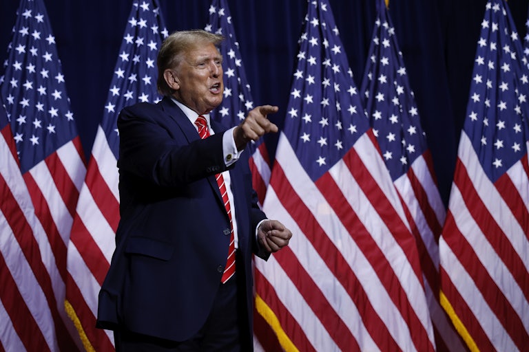 Donald Trump yells and points to something (presumably a crowd) in front of him. Several U.S. flags are behind him.