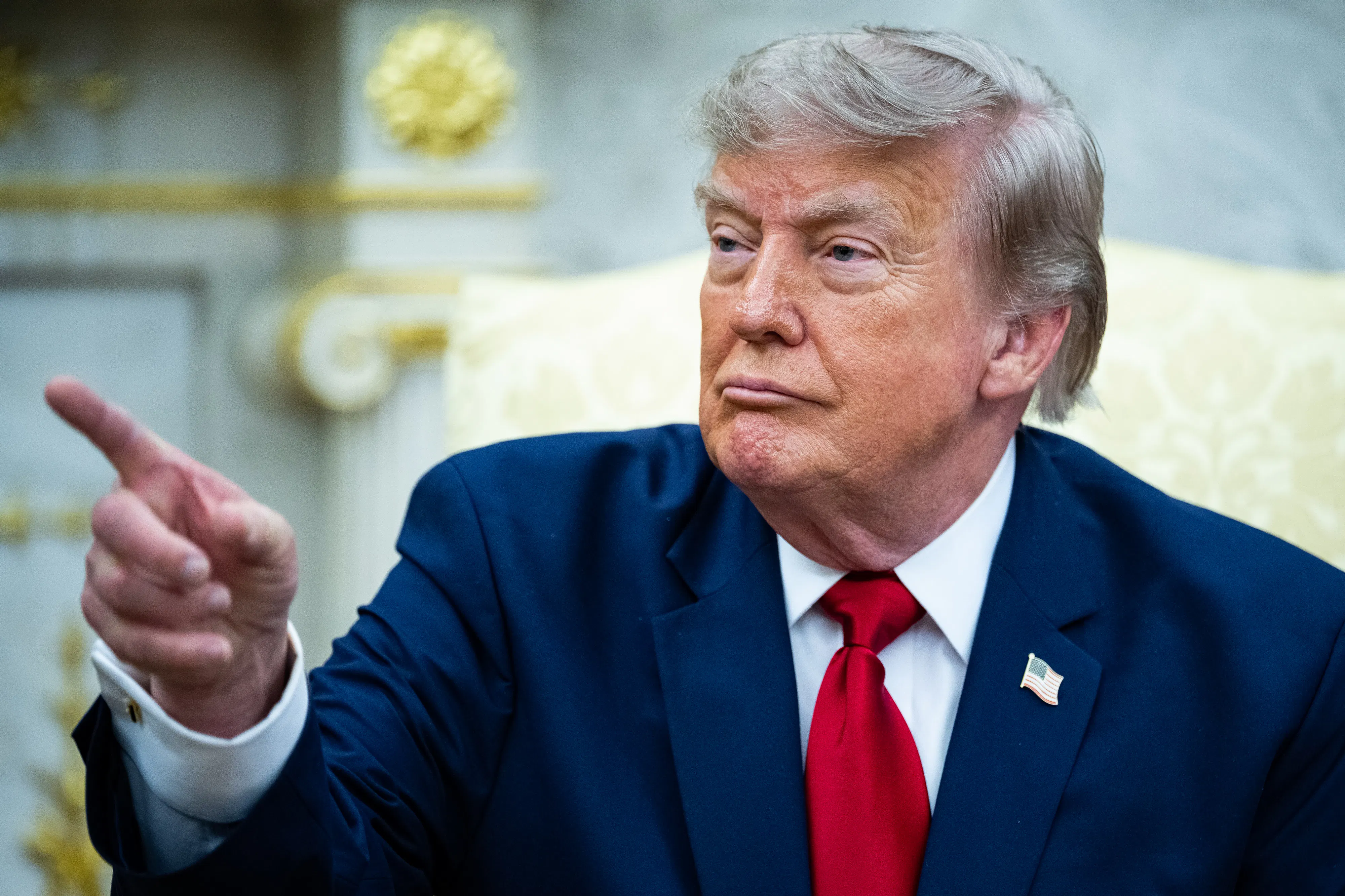 Donald Trump points at something while seated in the Oval Office of the White House.