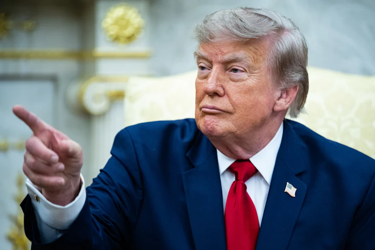 Donald Trump points at something while seated in the Oval Office of the White House.