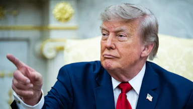 Donald Trump points at something while seated in the Oval Office of the White House.