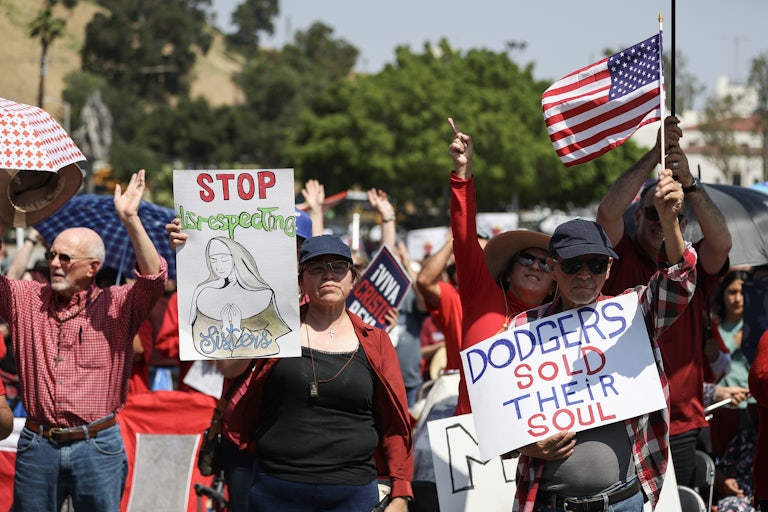 People hold signs and American flags at a protest, including a sign that says DODGERS SOLD THEIR SOUL