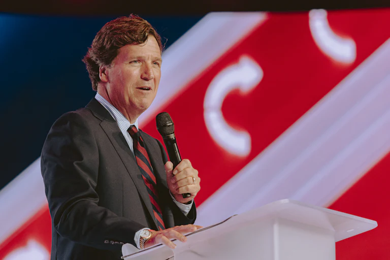Tucker Carlson holds up a microphone while speaking at a podium