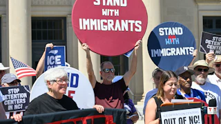 A crowd of people holding picket signs reading "Stand with Immigrants," "ICE Out," and "Stop Detention Centers in NC"
