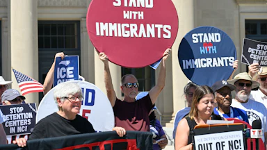 A crowd of people holding picket signs reading "Stand with Immigrants," "ICE Out," and "Stop Detention Centers in NC"