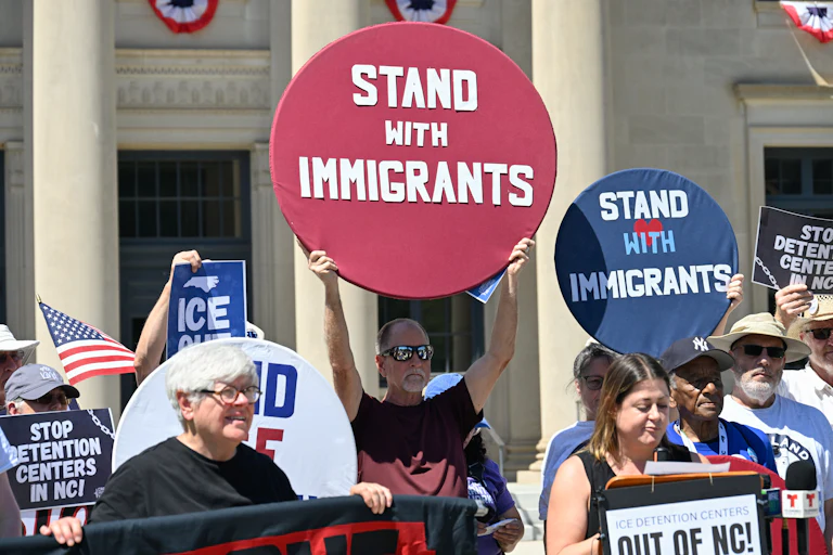 A crowd of people holding picket signs reading "Stand with Immigrants," "ICE Out," and "Stop Detention Centers in NC"