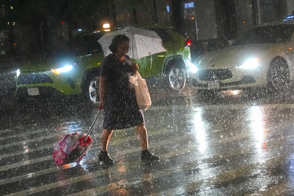A woman walks in heavy rain, pulling a cart, across a New York City crosswalk.