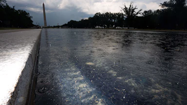 A low view of the Lincoln Memorial Reflecting Pool in the rain, with chipped paint and algae visible. In the background, the Washington Monument can be seen.