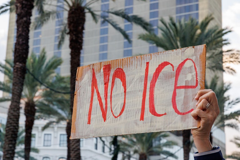 A hand holds a "No ICE" sign as palm trees are in the background