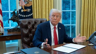 Donald Trump sits behind his desk in the Oval Office, preparing to sign something. He splays his hands outward as if to ask "what?"