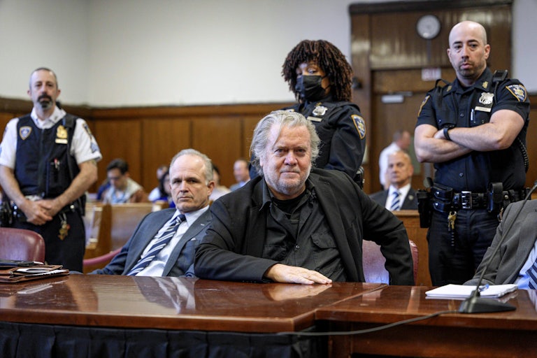 Steve Bannon sits at a courtroom table. Three security guards stand behind him.