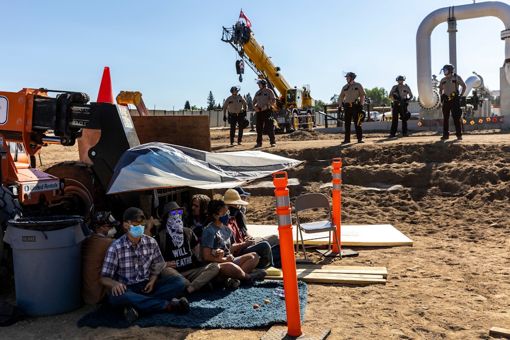 Activists sit beneath a tarp as law enforcement officers line up in front of construction equipment.