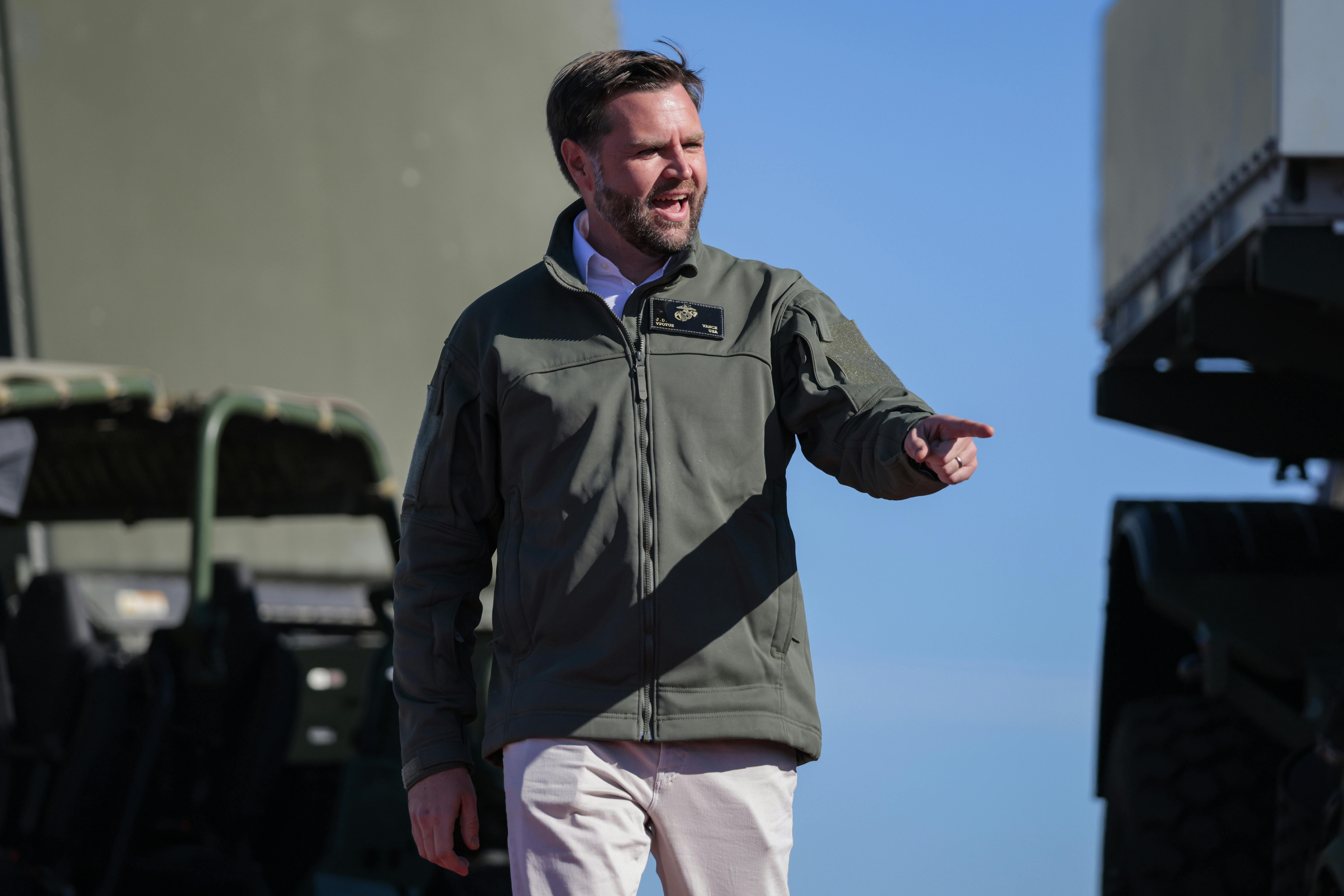 JD Vance smile sand points as a military vehicle is in the background.