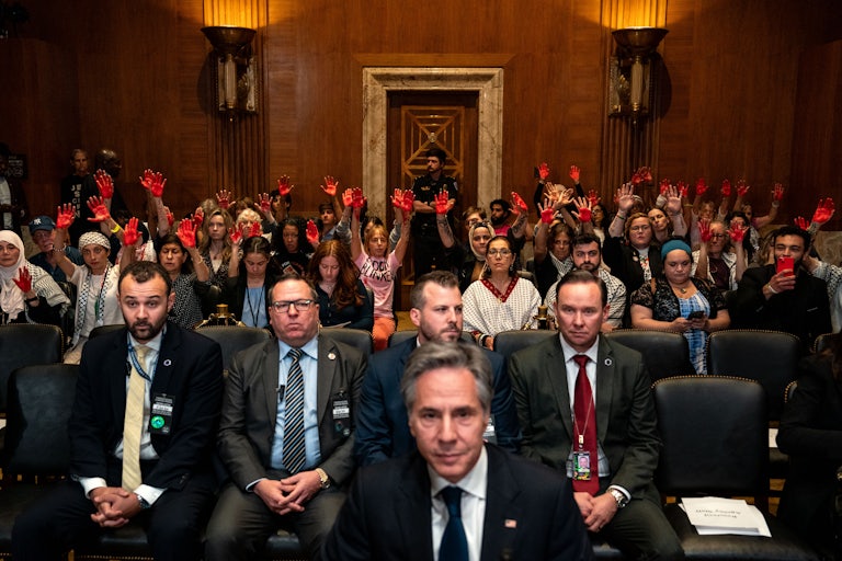 Antony Blinken is seated in the foreground. In the background are three rows of protesters raising their hands, painted red. Many are wearing keffiyehs.