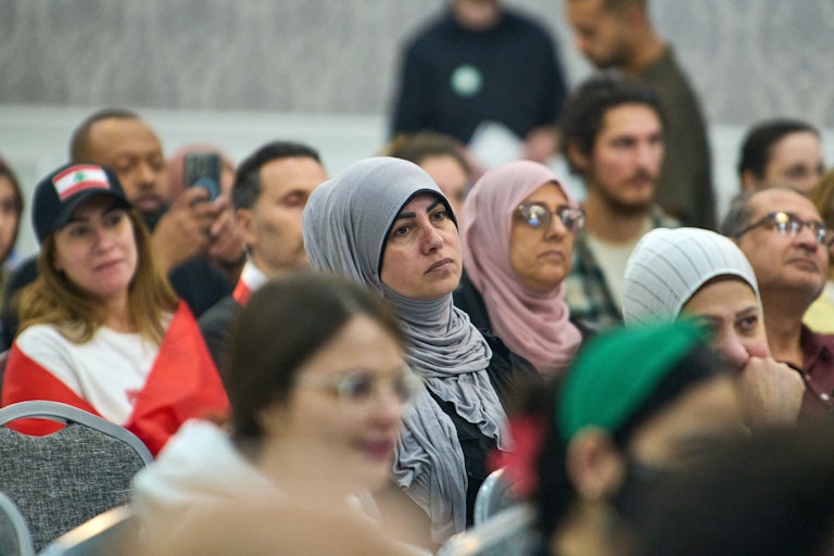 A group of hijabi women, non-hijabi women, and men (likely all Muslim, Arab, or brown) listen to a speaker (not pictured)..