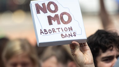 A hand holds a canvas sign in the air that reads "No MO Abortion Bans" with the outline of the state of Missouri. Other protesters are seen in background, out of focus.