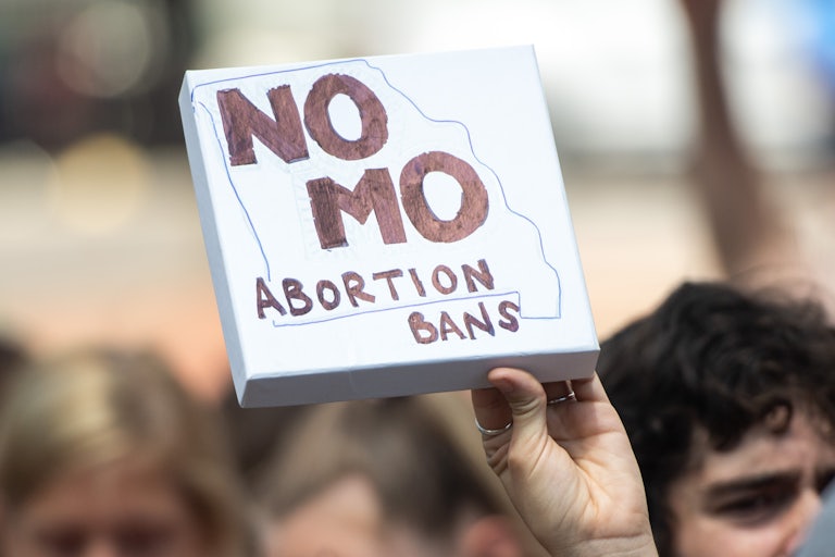 A hand holds a canvas sign in the air that reads "No MO Abortion Bans" with the outline of the state of Missouri. Other protesters are seen in background, out of focus.