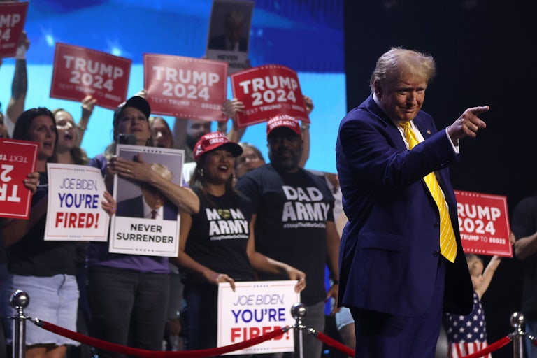 Donald Trump points and smiles as supporters behind him hold up "Trump 2024" and "Joe Biden You're Fired" signs