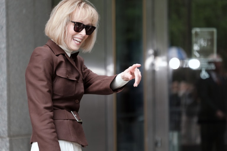E. Jean Carroll smiles and points to something off camera. She's wearing a brown coat and sunglasses.