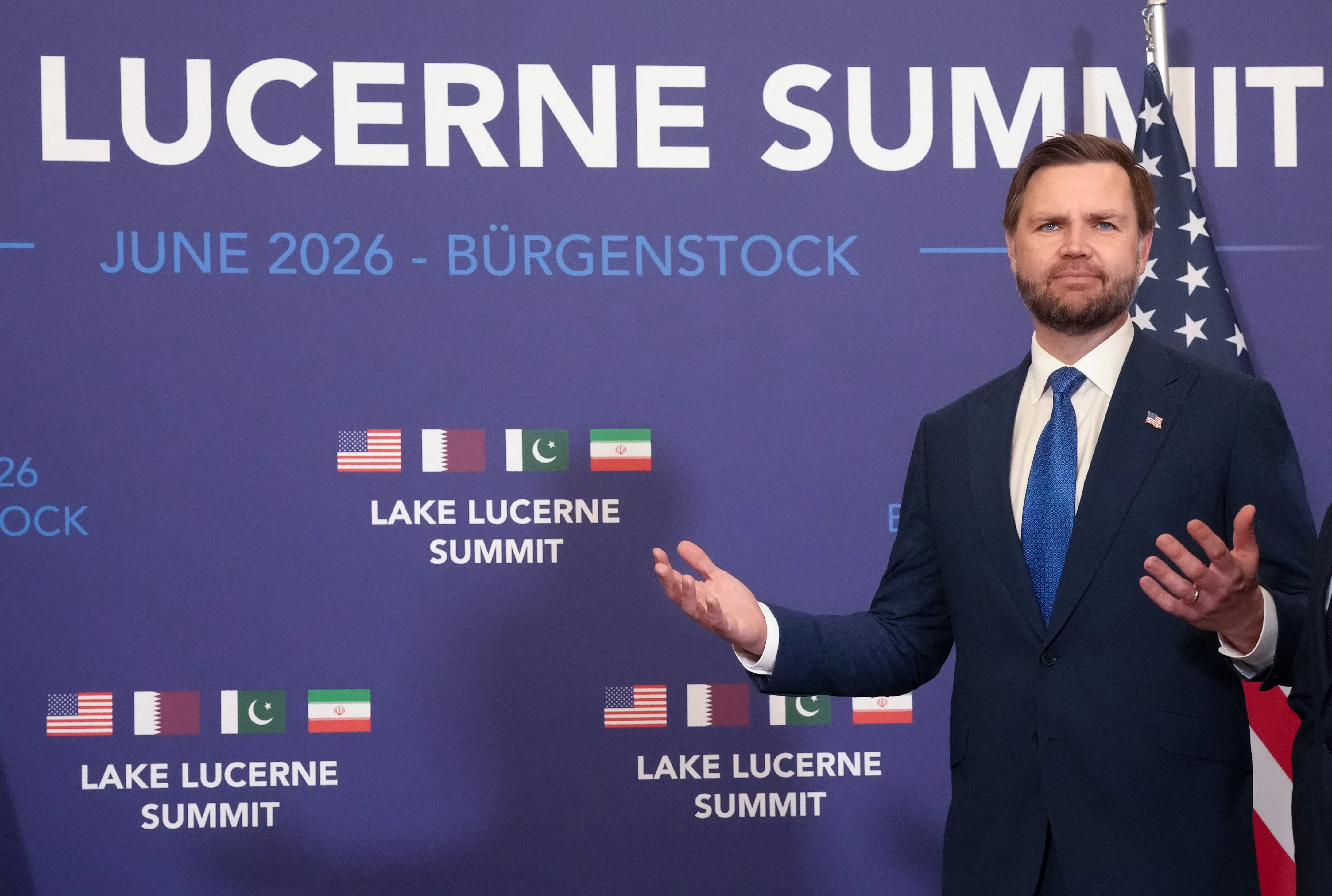 JD Vance stands in front a blue backdrop mentioning the Lucerne Summit on June 2016 in Switzerland with the flags of the U.S., Qatar, Pakistan, and Iran on the backdrop. 
