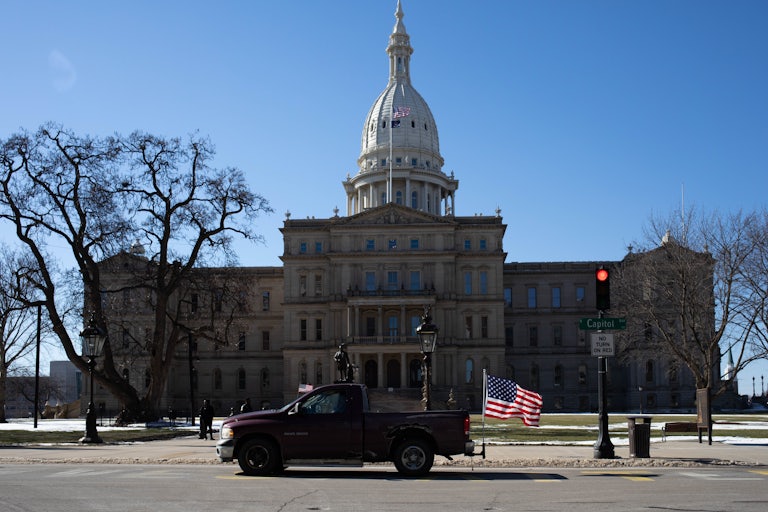 The Michigan state Capitol building. In front of it is a pick-up truck with an American flag flying off the end.