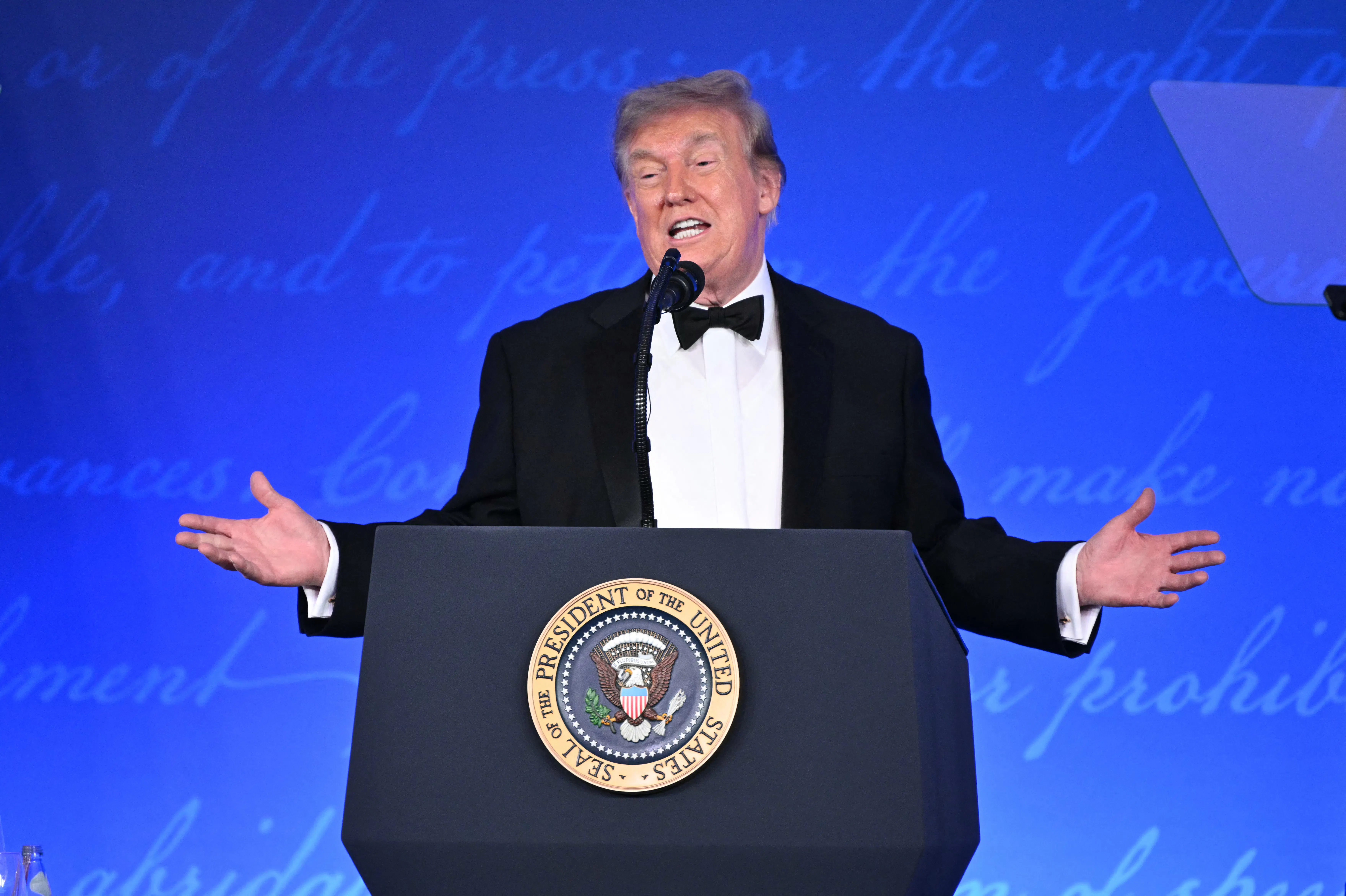 Donald Trump, wearing a tuxedo, spreads his hands while talking from a podium