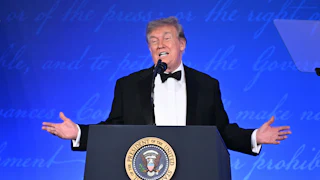 Donald Trump, wearing a tuxedo, spreads his hands while talking from a podium
