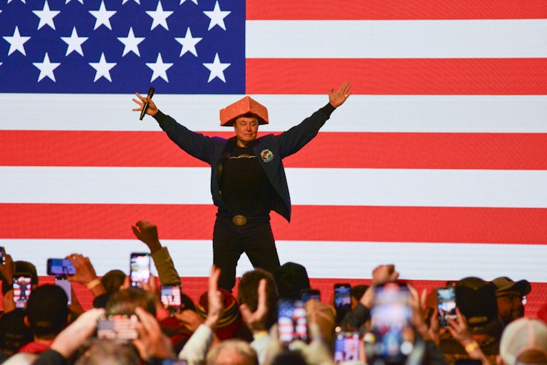 Elon Musk wears a cheesehead and raises his arms above his head while on stage in Wisconsin