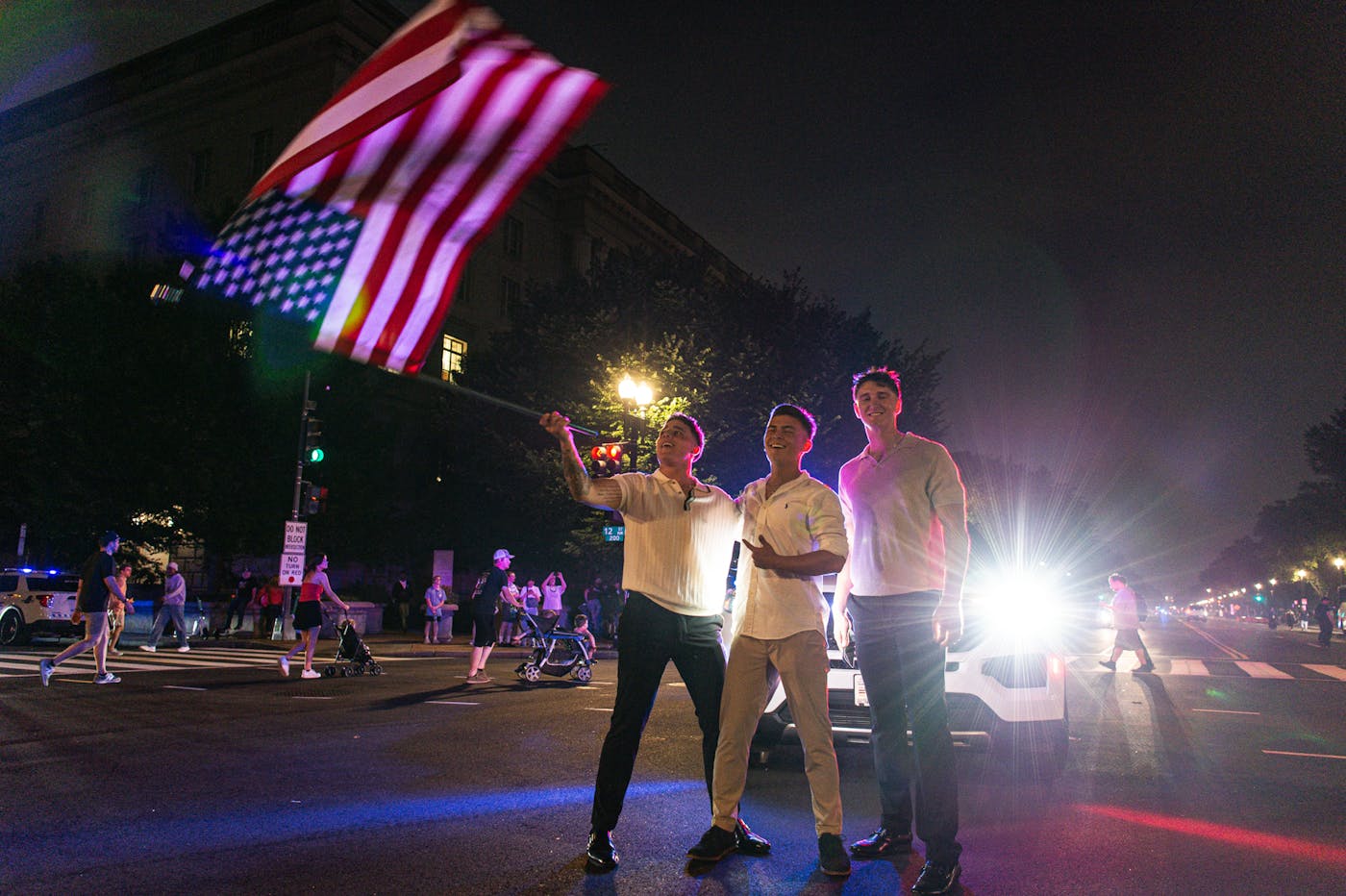 Three young men pose as one waves an American flag in front of flashing police lights in downtown Washington D.C. 