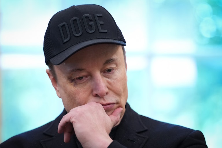 Elon Musk wears a black DOGE cap and puts his hand on his chin and stares downward as if he is deep in thought.