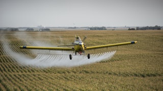 A crop duster flies over corn fields, trailing plumes of an unknown substance.