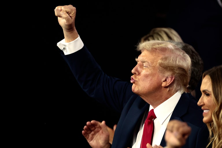 Donald Trump claps and blows a kiss (pursed lips). Someone beside him (face not seen) raises a fist in the air, as if in victory.