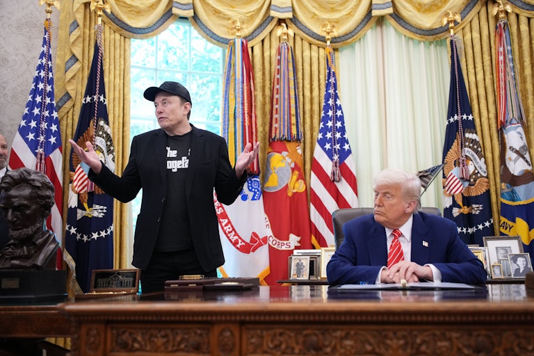 Elon Musk shrugs while standing next to Donald Trump, who sits at his desk in the Oval Office