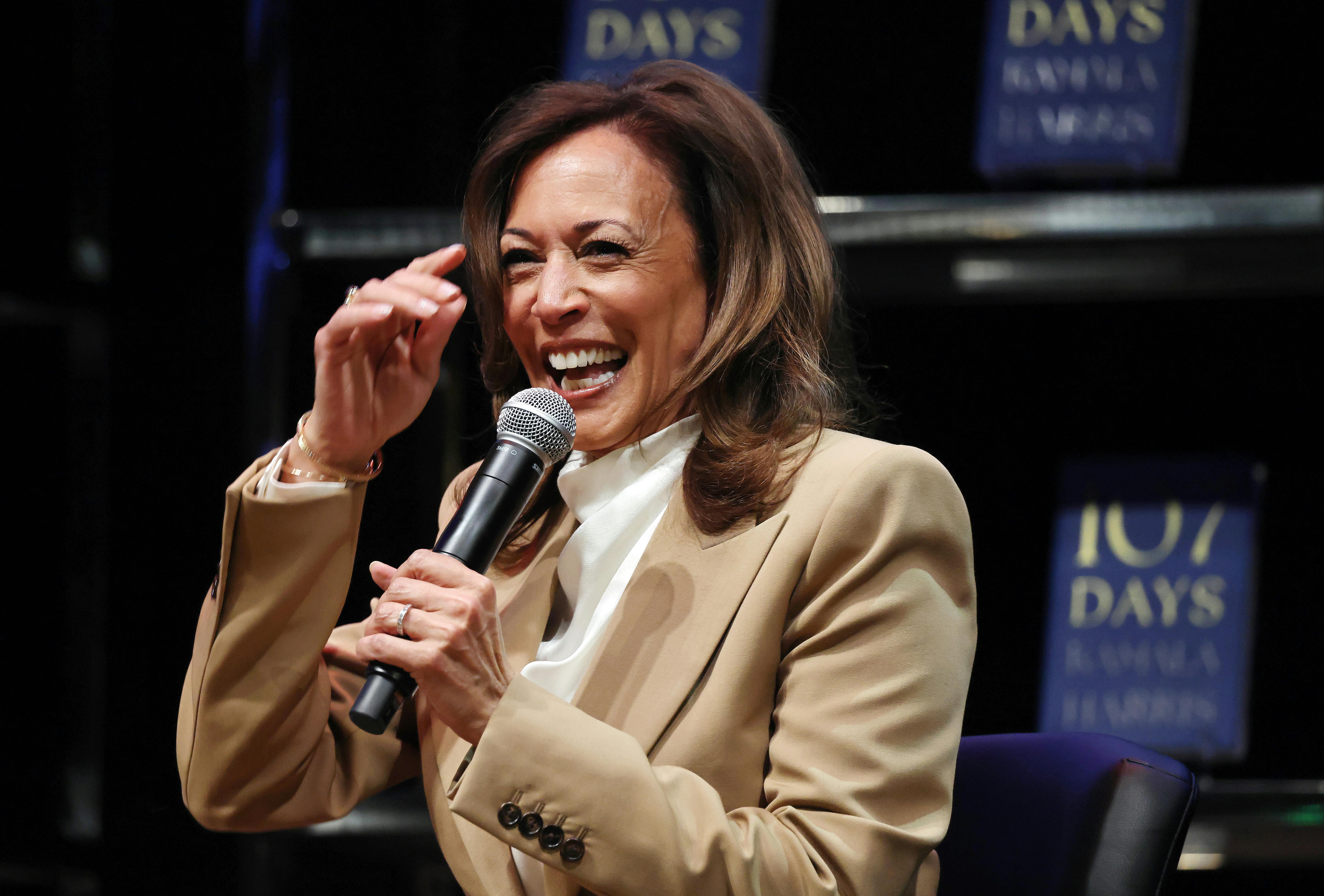Kamala Harris laughs while she speaks on a mic. Her book 107 Days is in the background.