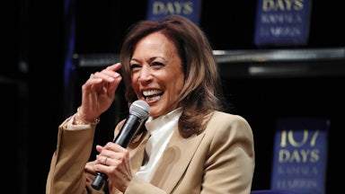 Kamala Harris laughs while she speaks on a mic. Her book 107 Days is in the background.