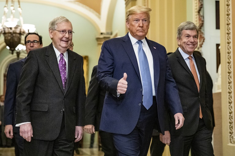Mitch McConnell smiles and follows Donald Trump who holds up a thumbs up for the camera. Other men surround them.